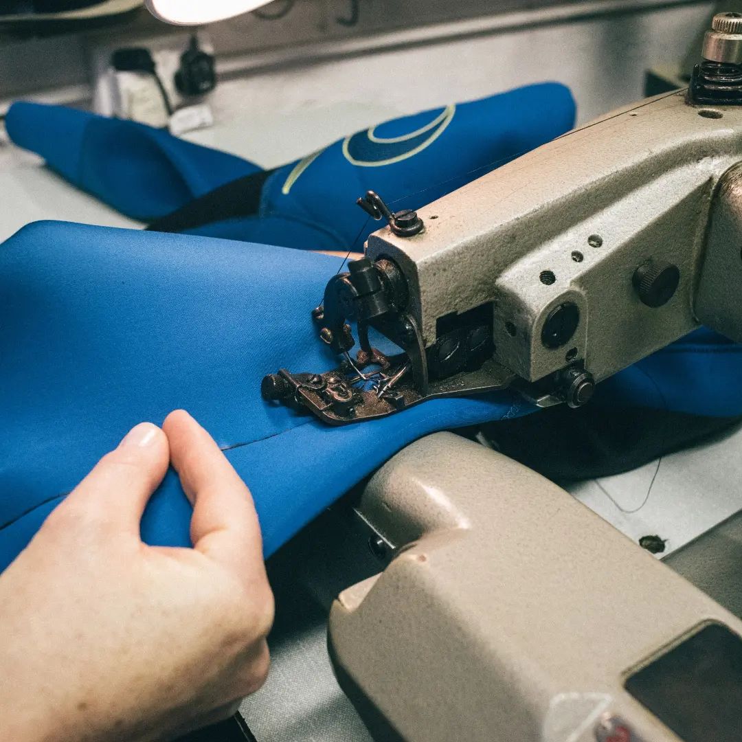 Close-up of hands feeding blue neoprene through an industrial sewing machine during a wetsuit repair