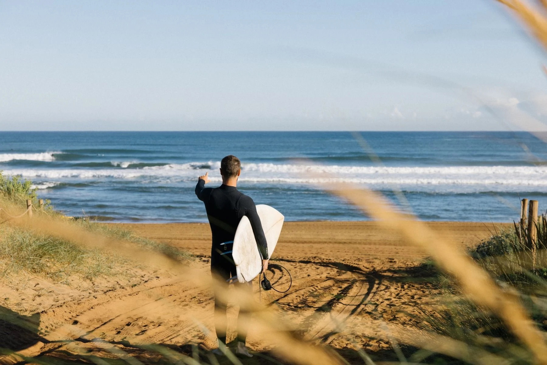 Surfer in a black wetsuit walking down a sandy dune path to the beach with a board under one arm