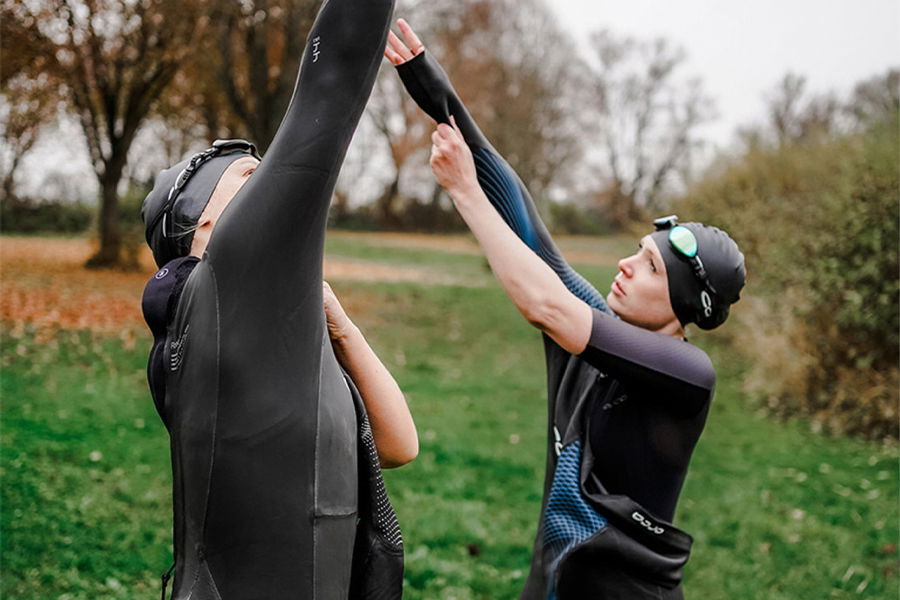 Two swimmers in wetsuits and caps on grass, one helping the other peel off a wetsuit after a swim