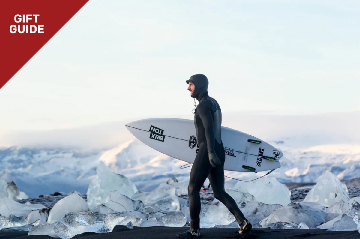 Surfer im Winter-Neoprenanzug mit Kopfhaube trägt sein Surfboard durch die Dünen
