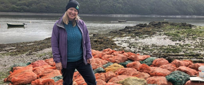 Woman in a purple jacket and beanie standing on lichen-covered rocks beside an estuary at low tide