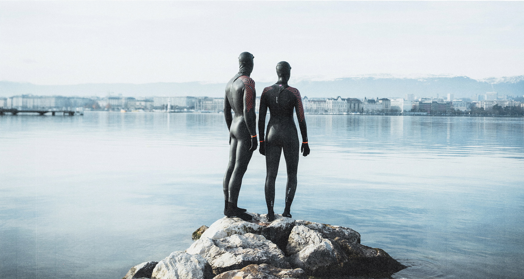 Two swimmers in wetsuits standing on rocks looking out over a flat, cold sea towards a town