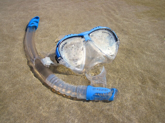 Clear and blue snorkelling mask with attached snorkel lying on damp sand